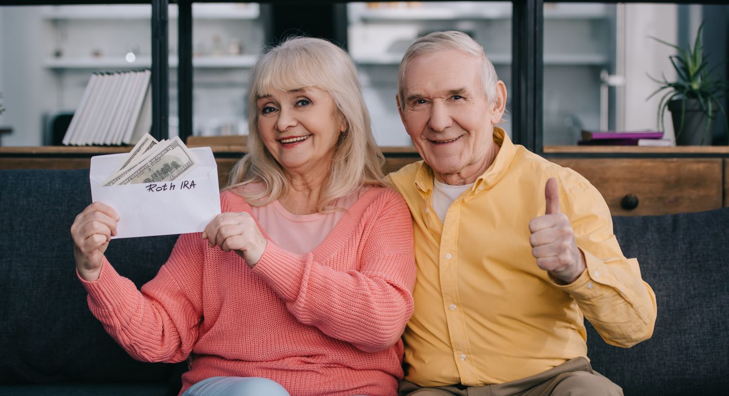 Senior couple showing thumb up sign while holding envelope with 'roth ira' lettering and dollar.
